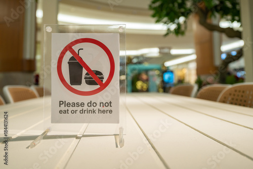 A clear safety sign on a white table in a busy indoor public space, reminding visitors do not to eat or drink. The sign is framed and features a red prohibition