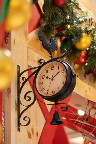 A charming vintage clock mounted on a wooden wall, featuring a rooster weather vane and a hanging bell. Surrounded by festive Christmas decorations and rustic decor creating a cozy holiday.