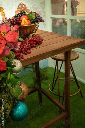 A festive indoor table centerpiece featuring a wood table and chair, a bowl of colourful fruit, lush greens, and bright poinsettias.