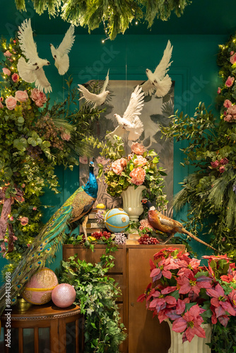 Vibrant indoor garden-inspired display featuring doves in flight, peacocks perched, and a lush arrangement of pink flowers, greenery, and decorative elements set against a teal backdrop for festive.