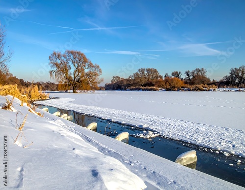 Snowy river landscape under a clear blue sky, with trees and a bank covered in snow