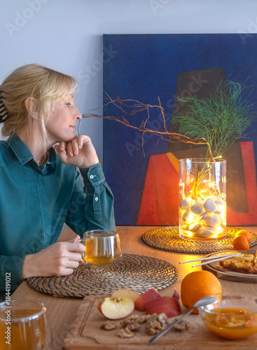 A woman meditatively looks at the magical light of a garland decorating a glass vase with ikebana, sitting at a table with a cup of tea in her hand during a tea party