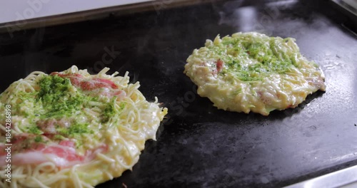 Noodle topped okonomiyaki with bacon sizzling on a heated teppan in Japan as the camera tracks gently across, showing drifting steam and early cooking texture in the scene