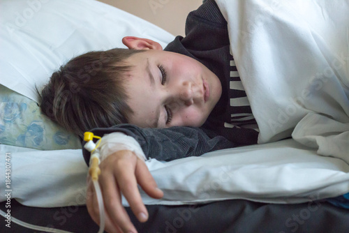 A sick preschool-aged boy is lying in a hospital bed. A venous catheter is visible on his arm.