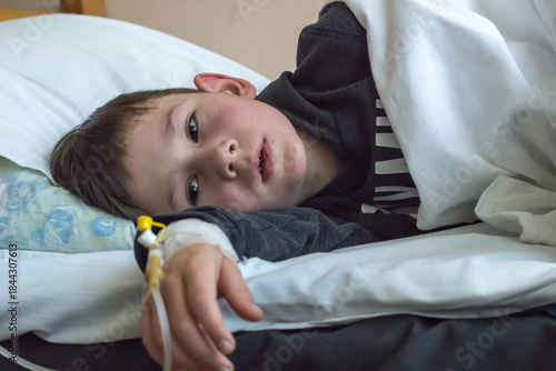 A sick preschool-aged boy is lying in a hospital bed. A venous catheter is visible on his arm.