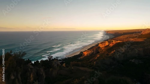 Majestic ocean cliffs bathed in warm sunset hues stretch along the coastline. the expansive view captures gentle waves meeting rugged cliffs under a serene sky, offering a tranquil natural scene perfe