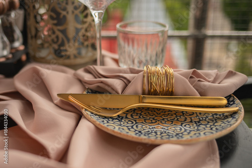 Sophisticated table setting featuring gold cutlery, a patterned ceramic plate, and a dusty rose fabric napkin with a gold ring, set outdoors for a luxury event or celebration.