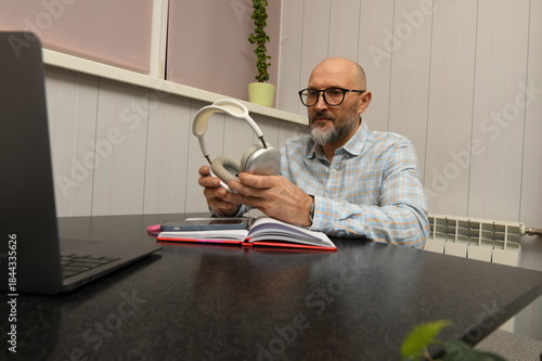 A man sits at a desk in an indoor space, holding headphones while looking at a laptop. A notebook and pen are nearby as he prepares for a work task