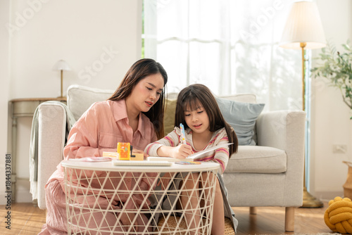 Asian Family, mother teaching her daughter in the living room while doing homework. Study, drawing, and writing in book with parents helping their girl kid at home. Children education concept
