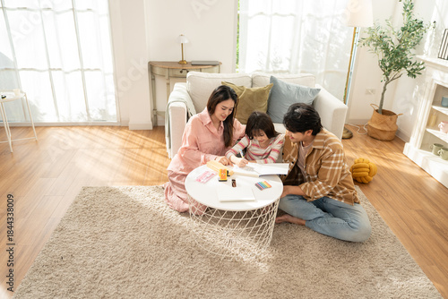 Asian Family, father and mother teaching their daughter in the living room while doing homework. Study, drawing, and writing in book with parents helping their girl kid at home.