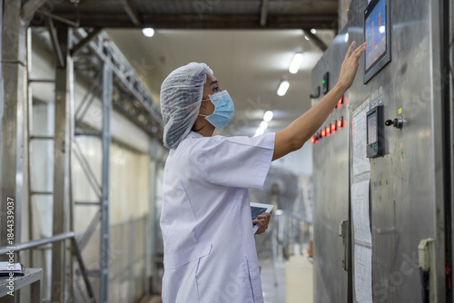 Female inspector wearing protective gear and hairnet examines canned fish production quality, checks Industrial pressure cooker, monitors food processing , and records data using tablet.
