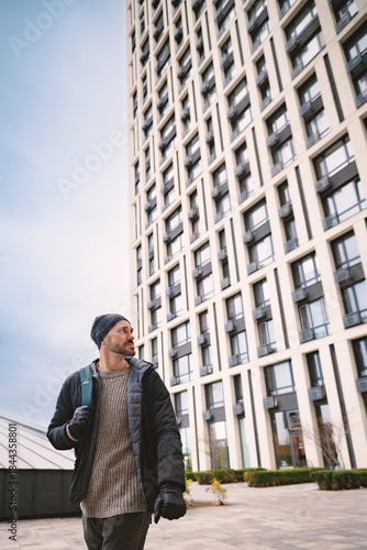 Wallpaper Mural Young man in a winter jacket and beanie walking past a modern high-rise building on a cloudy day, looking thoughtfully to the side. Urban lifestyle concept. Torontodigital.ca