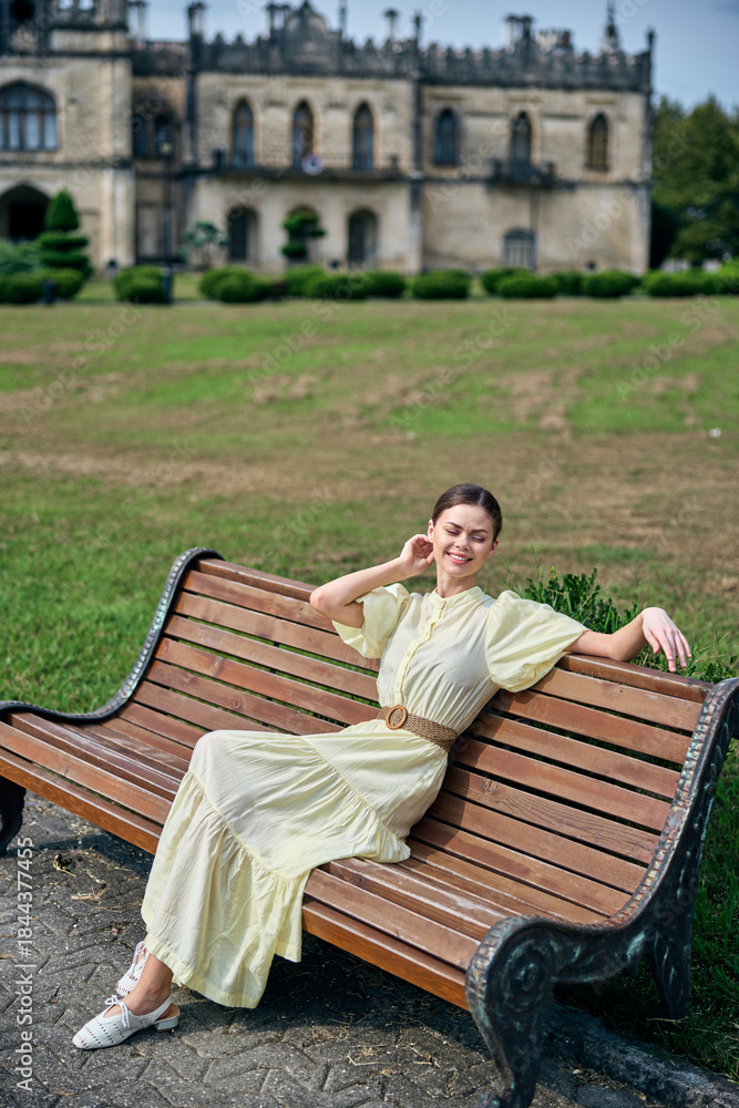 Fototapeta premium Elegant woman in a pale dress relaxing on a wooden bench in a park with a historic castle in the background, creating a calm portrait, soft light enhances fabric, poised posture, tranquil atmosphere,