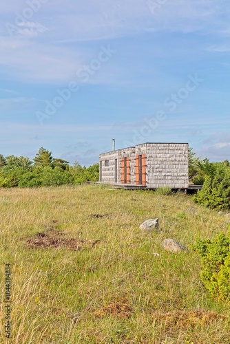 Small, shingle-clad modern off-grid cabin or retreat in grassy and shrubs surrounding in summer with blue sky with light clouds, Türju, Saaremaa, Estonia.