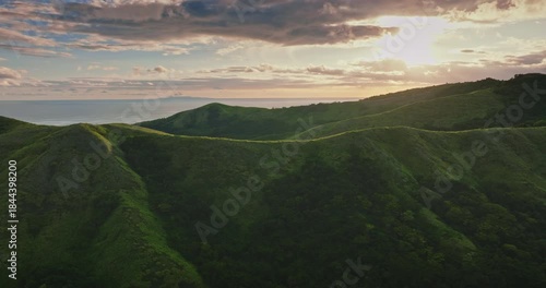 Lush green hills and valleys creating a beautiful landscape in Fiji, with the Pacific Ocean visible in the distance under a dramatic sky with golden sunset light