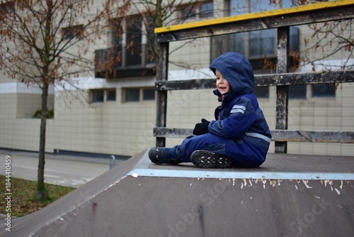 Toddler boy sitting on top of skate ramp in urban playground during autumn. Child wearing warm winter clothes enjoying calm outdoor moment, building confidence and independence in early childhood.