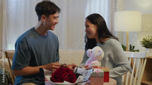 Couple young lovers, faces happy on valentine day at home, share romantic love with red roses, gifts, teddy bear, celebrating special occasion together.