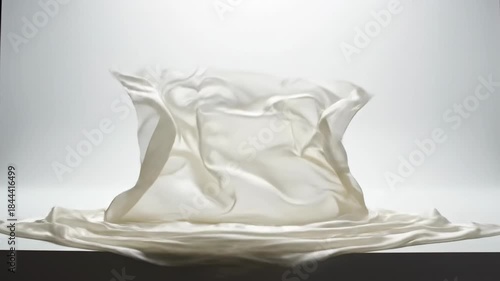 Elegant slow motion shot of lightweight sheer ivory silk fabric gracefully billowing and settling onto a clean minimalist white studio platform product display, smooth, minimalist