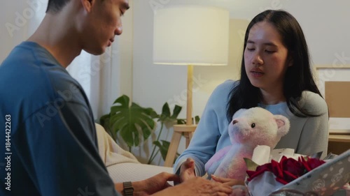 Love connects happy couple, man holding hands of laughing woman, celebrating valentine day at home, romantic moment with soft teddy bear and beautiful red roses.
