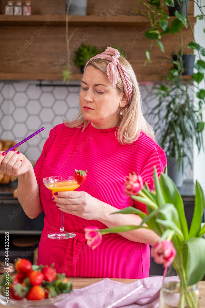 Fototapeta premium Woman in bright pink dress preparing a colorful cocktail in a modern kitchen, surrounded by fresh flowers and fruits, showcasing vibrant culinary creativity and style