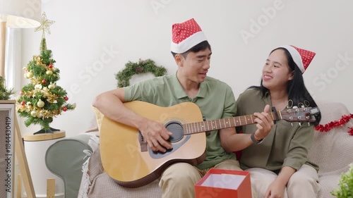 Happy young couple enjoying christmas holiday at home, people wearing Santa hats, man playing guitar for woman on couch, xmas tree brightly decorated, presents waiting, creating festive family memory.