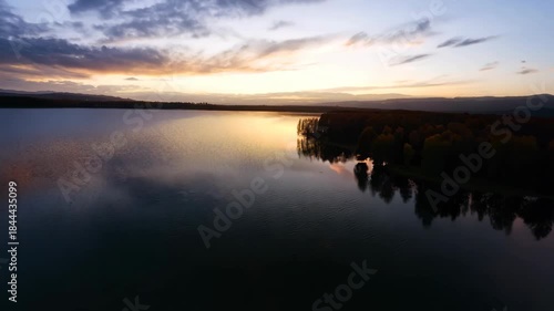 Scenic view of a tranquil lake at sunset, featuring calm reflective waters and silhouetted autumn trees under a dramatic sky. ideal for designs focusing on nature, tranquility, and serene landscapes.