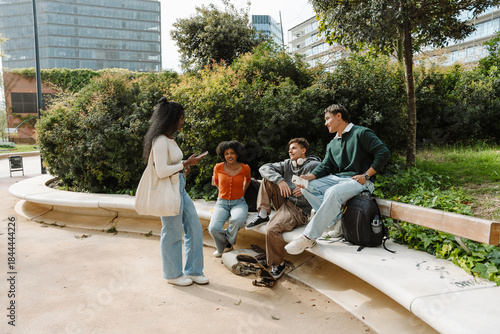 A group of three students are sitting on a stone bench and talking to a female student standing across from them holding a phone