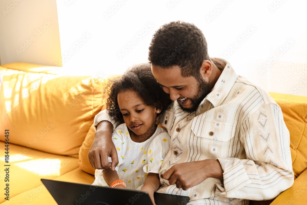 Obraz premium Father laughing and holding his arm around his daughter while they look at a laptop and sit on the couch