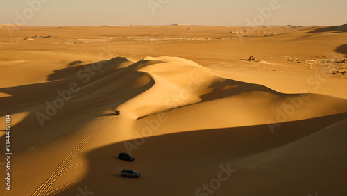 Several off-road vehicles drive across the dunes. Wide desert landscape with rolling golden sand dunes and long dramatic shadows cast by low sunlight. Western Desert near Siwa oasis, Egypt