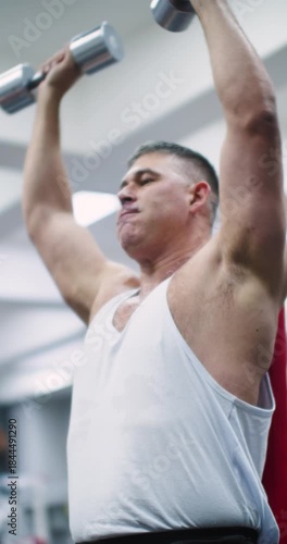 Seated bench press with dumbbells for the deltoid muscles. Deltoid workout with dumbbells. Cropped vertical 4k footage of a middle aged brutal Caucasian man during gym workout.