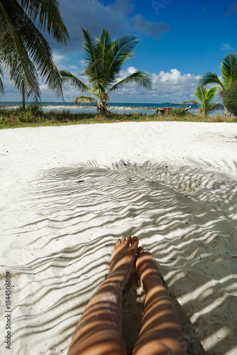 Woman relaxing on tropical beach under palm trees. A bright closeup showing calm and peaceful vibes. Vacation lifestyle, summer vibes, serene escape. Woman legs with coconut leaf shadow.