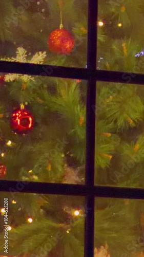 Vertical video. Close Up of Christmas Tree behind Window showing Bright Red Ornaments and Twinkling Lights creating Cozy Holiday Mood in Soft Angle View, Festive Interior Concept