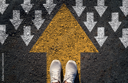 Top view of shoes standing on a bold yellow arrow pointing forward against a field of white arrows pointing downward symbolizing disruption leadership innovation and moving in a different direction
