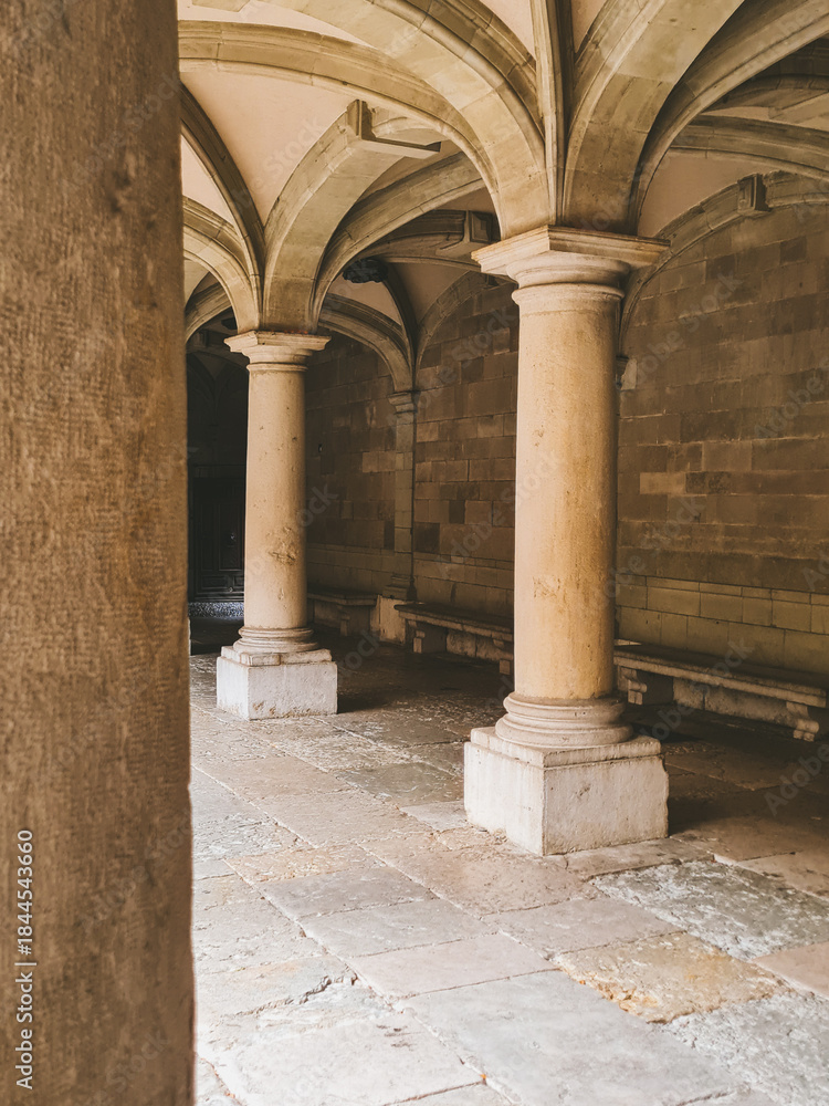 Fototapeta premium An arched gallery with massive stone columns. A bright stone space with symmetrical vaults, soft natural light, and textured stonework.