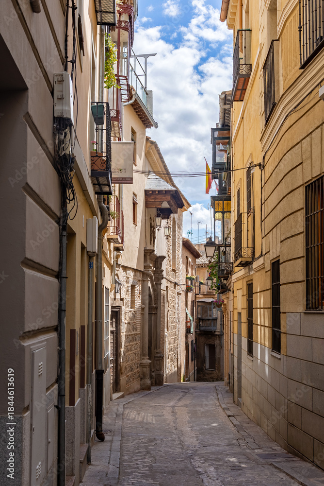 Naklejka premium Vertical view down a narrow cobblestone Toledo street, lined with warm stucco facades, wrought-iron balconies, exposed stone, and a covered wooden balcony in the distance