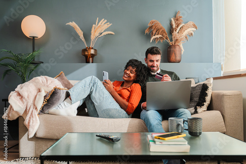 A couple happily engaged in online shopping, using a laptop and smartphone to browse products while enjoying their time together on the couch in their cozy living space.