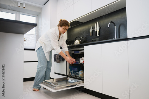 Young woman placing cups and dishes into an open dishwasher in a modern minimalist kitchen. Casual domestic routine, standing pose, contemporary interior, natural indoor daylight.