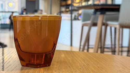 Flat white coffee beverage in transparent orange-tinted, faceted glass cup on wooden café table with blurred interior in background