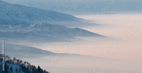 Snow-capped mountains covered with layers of fog or smog, with trees in the foreground. Winter landscape with soft light and cold atmosphere.