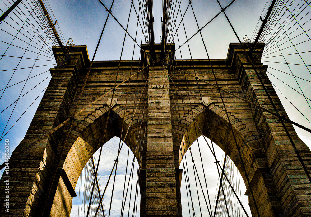 Fototapeta premium Dramatic architectural view of the Brooklyn Bridge, captured from below with strong lines, stone arches and converging cables creating a powerful geometric composition.