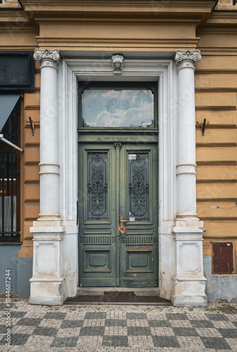 Historic building entrance with weathered green double doors framed by classical white columns and ornate stone details.