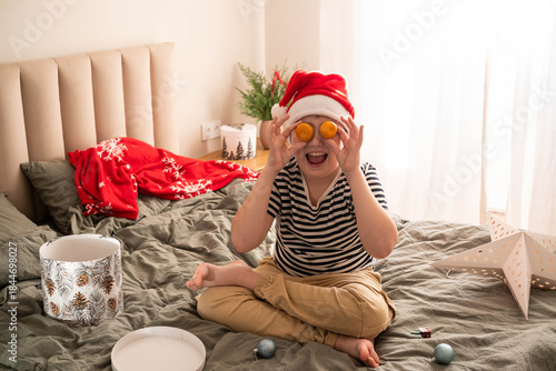 Playful child in a Santa hat sits on a bed holding tangerines over eyes and laughing. Cozy home interior, natural light, festive winter holidays, joyful childhood moment.