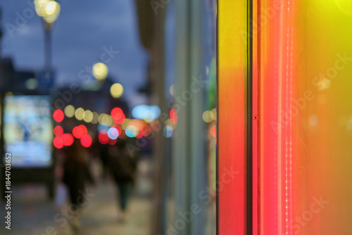 Street at dusk or night, focusing on the vibrant red and yellow neon lights of a storefront contrasted with a blurred street scene in the background. selective focus
