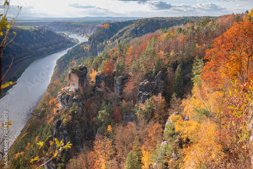 Saxon Switzerland, Bastei nature reserve. Wide Panoramic a vibrant autumn forest surrounded by sunlight, river, mountains on the horizon, and a cloudy sky