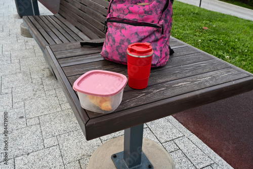 Lunch break with food container and drink on a wooden table in a park during daylight hours