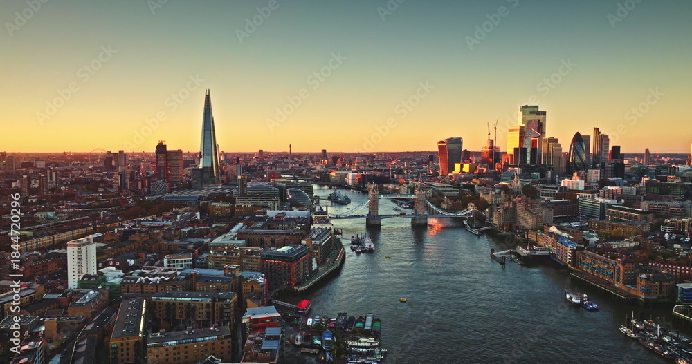 Fototapeta premium England, aerial London skyline at sunset with iconic landmarks Tower Bridge, The Shard skyscraper, and illuminated buildings of Canary Wharf on River Thames. Modern cityscape under bright evening sky