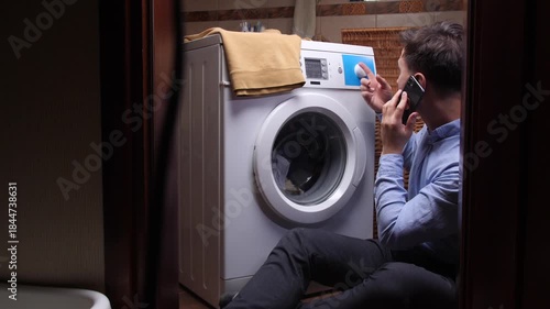 Man sitting on the floor staring into a broken front load washing machine, talking on his phone while frustrated and calling for repair or troubleshooting help in his apartment