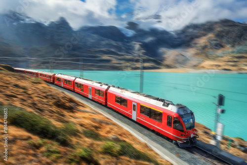 Modern red train is moving along railway in swiss alps mountain range near lake on sunny day with cloudy sky in autumn. Landscape. Bernina Express. High speed train with motion blur effect. Travel