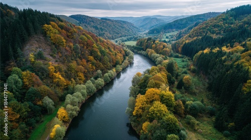 A river flows through a valley surrounded by hills and forests. Trees display autumn colors of orange and yellow. Clouds cover the sky as sunlight filters through casting shadows.