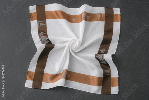 A white cotton handkerchief with a wide brown and tan border, featuring a sailboat pattern is folded into a swirl pattern and placed on a dark gray surface.